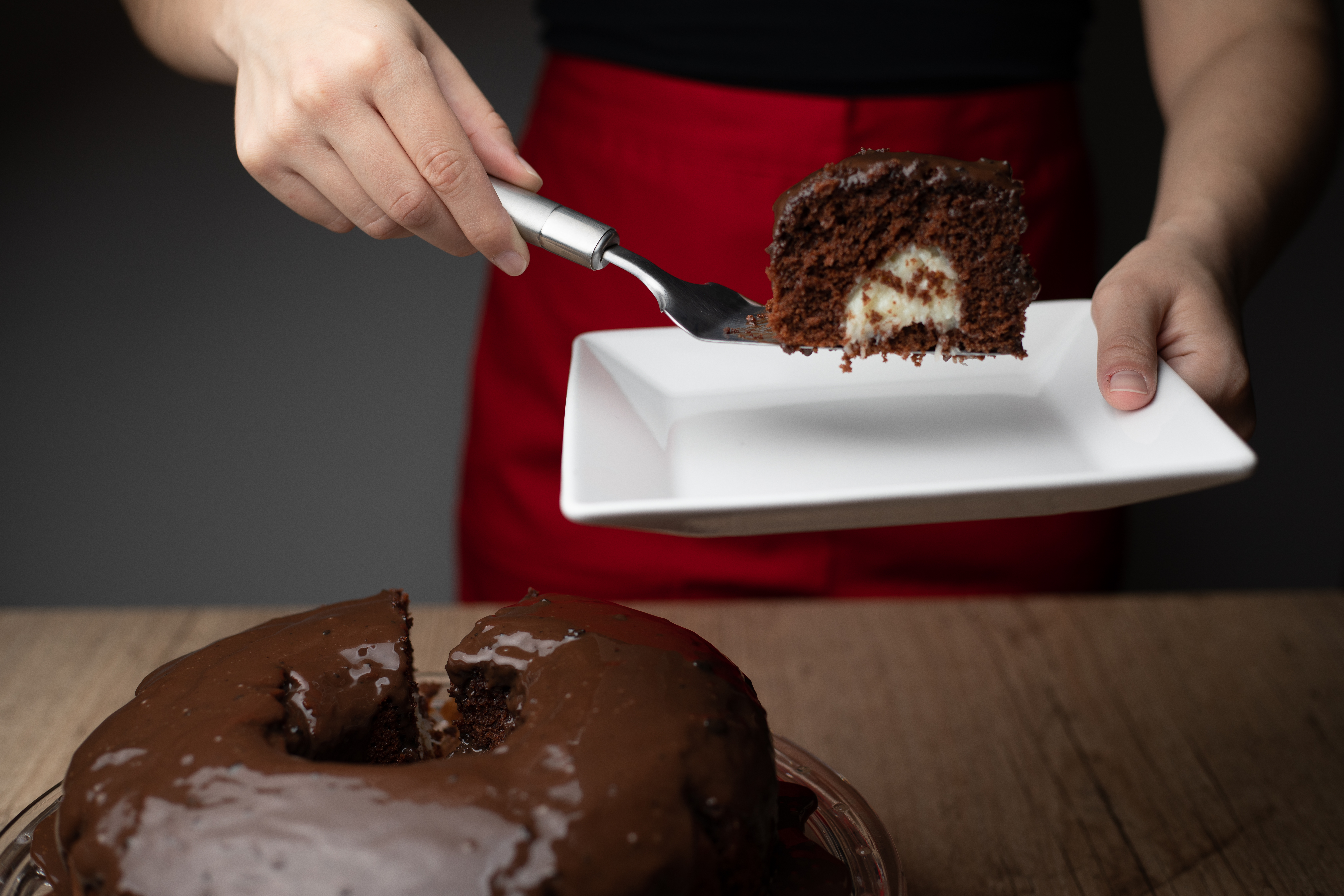 Bolo de chocolate com furo no meio coberto de brigadeiro à esquerda inferior da foto. À direita superior, pessoa com avental vermelho segurando prato e espátula com pedaço de bolo de chocolate com recheio de cocada ao centro. 